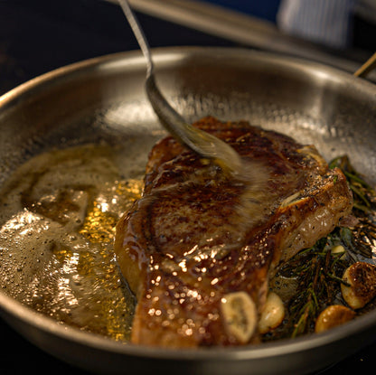 Searing steak in a pan with herbs and garlic, close-up view.