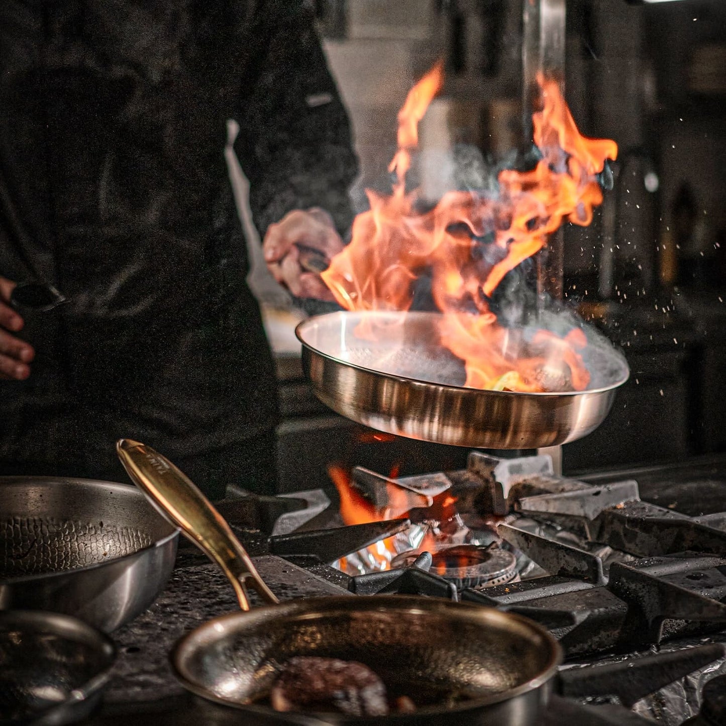 Person cooking over a flaming stove with multiple Titanium pans on a stovetop.